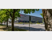 Außenansicht der S-Arena Bernau, einer modernen Veranstaltungshalle mit einer dunklen, geometrisch gemusterten Fassade. Im Vordergrund sind eine Straße, Bäume und ein Gehweg zu sehen, umgeben von Grünflächen und Verkehrsschildern.