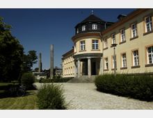 Gelbes historisches Gebäude mit Rundturm und Säulenportal, umgeben von gepflegten Hecken und einem gepflasterten Weg; im Hintergrund sind industrielle Schornsteine und Bäume sichtbar.