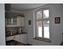 Helle Küche mit weißen Einbauschränken, Holzarbeitsplatten und einem großen, weiß gerahmten Fenster, das den Blick auf eine verschneite Außenlandschaft freigibt. Eine Dunstabzugshaube, kleinere Dekorationsbilder und ein Heizkörper unterhalb des Fensters ergänzen die Einrichtung.