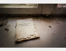 Ein altes, abgenutztes Heft mit der Aufschrift „Fahrtenauftrags- und -nachweisbuch“ liegt auf einer staubigen Fensterbank, umgeben von Schmutz und Spinnweben.