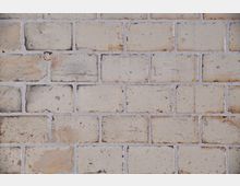 Nahaufnahme einer hellbeigen gemauerten Ziegelwand mit leicht unregelmäßiger Oberfläche und sichtbaren Fugen. Die Ziegel zeigen Gebrauchsspuren wie Verfärbungen und Kratzer.