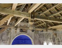 Holzgebälk einer historischen Dachkonstruktion mit sichtbaren Balken und Ziegeln, darunter eine bemalte, blaugoldene Apsiskuppel und teils freigelegte Mauerwerkselemente. Die Umgebung zeigt Spuren von Verwitterung und Restaurierungsbedarf.