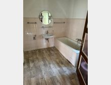 Ein Badezimmer mit einer Badewanne, einem Waschbecken und einem runden Spiegel mit zwei Wandlampen darüber. Die Wände sind mit beigen Fliesen verkleidet, der Boden besteht aus Holzoptik-Vinyl, und es gibt eine Holztür im Vordergrund.