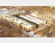 Luftaufnahme eines verlassenen Industriekomplexes mit mehreren langgestreckten Gebäuden und flachen, schneebedeckten Dächern, aus denen kleine Bäume und Sträucher wachsen. Die Anlage liegt am Waldrand in einer winterlichen Landschaft, umgeben von kahlen Bäumen und verschneiten Wegen.