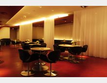 Ein eleganter Innenbereich mit roten Böden, weißen Vorhängen und indirektem, warmem Licht. Die Einrichtung umfasst runde Tische mit schwarzen Polsterstühlen und kleinen Pflanzendekorationen auf jedem Tisch.