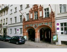 Fassade eines historischen Gebäudes in Berlin mit roten Backsteinelementen und gotisch inspirierten Details. Über den drei Rundbögen steht der Schriftzug „Berliner Handwerker-Verein“, seitlich sind Wohnhäuser und parkende Autos zu sehen.