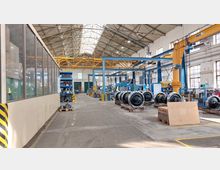 Industriehalle mit hoher Decke und Oberlichtern, ausgestattet mit gelben Krananlagen und blauen Arbeitsgestellen. Im Vordergrund liegen Zugräder auf Paletten, links ist ein Raum mit Glaswänden abgetrennt.