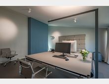 Ein modernes Büro mit einem großen Schreibtisch, darauf zwei Monitore, eine Tastatur, eine Maus und ein Blumenstrauß in einer Glasvase. Im Hintergrund sind eine blaue Wand, ein Aktenschrank und eine Stehlampe zu sehen; links steht ein grauer Bürostuhl.