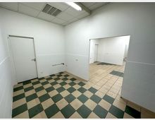 Innenraum mit weißen Wänden und gefliestem Boden in grün-beigen Karomustern, ergänzt durch eine Zwischentür zu einem benachbarten Raum. Die Decke hat weiße Platten mit integrierten Leuchtstofflampen und Lüftungsgitter.