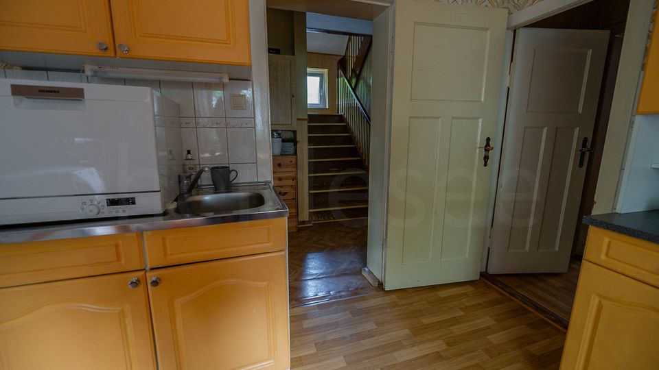 Blick in eine kleine Küche mit gelben Holzschränken, Geschirrspüler und Spüle vor einer gefliesten Rückwand. Durch eine offene Tür ist ein Flur mit Holztreppe und einem Fenster am Ende zu sehen.