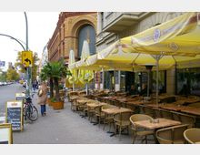 Straßencafé mit gelben Sonnenschirmen und vielen leeren Tischen und Stühlen aus Rattan entlang eines Gehwegs neben einem historischen Backsteingebäude. Im Hintergrund sind Fußgänger, geparkte Fahrräder und eine Straßenlaterne zu sehen.
