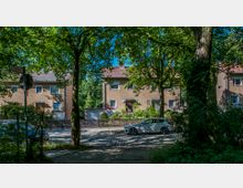 Reihenhäuser mit brauner Fassade und roten Ziegeldächern stehen hinter einem Gartenzaun, umgeben von grünen Bäumen. Im Vordergrund führt eine gepflasterte Straße vorbei, auf der ein weißes Auto geparkt ist.