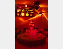 Ein dimm beleuchteter Raum in Rot- und Orangetönen, mit einer zentralen achteckigen Sitzgelegenheit, auf der eine Vase mit Blumen steht. Die Decke ist mit reflektierenden Elementen und kreisförmigen Leuchten dekoriert, die einen Retro-Stil vermitteln.