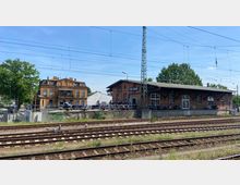 Ehemaliger Backsteinbahnhof mit zweigeschossigem Hauptgebäude und angeschlossenem Güterschuppen, genutzt als Harley-Davidson-Händler. Im Vordergrund befinden sich mehrere Bahngleise, im Hintergrund Bäume und blauer Himmel.