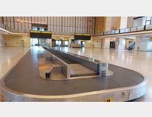 Großer, leerer Gepäckausgabebereich in einem Flughafen mit einem zentralen Gepäckband, hohen Wänden aus Stein und Glas und einer Beschilderung mit gelben Informationsanzeigen. Im Hintergrund befinden sich Türen zum Ausgang, ein Restaurant-Schild und Treppen zu einer oberen Ebene.