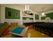 Ein großer, klassisch gestalteter Saal mit grünen Wandflächen, weißen Holzvertäfelungen und vergoldeten Kronleuchtern. Im Vordergrund ein Rednerpult, im Hintergrund Sitzreihen, Holzpodeste und gerahmte Porträts an den Wänden.