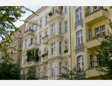 Mehrstöckige Wohnhäuser im Altbaustil mit verzierten Fassaden und Balkonen, teils mit Blumenschmuck. Im Vordergrund sind Bäume zu sehen, die teilweise die Gebäude verdecken.