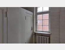 Ein einfacher Innenraum mit weißen Fliesenwänden, einem Fenster mit Sprossenteilung und einem Blick auf eine rote Backsteinfassade. Ein Heizkörper befindet sich unter dem Fenster, daneben eine geschlossene Zimmertür.