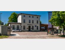 Vorderansicht eines kleinen Bahnhofsgebäudes mit weißer Fassade und der Aufschrift „Rheinsberg (Mark)“, davor ein gepflasterter Vorplatz. Rechts stehen Informationsvitrinen und ein Fahnenmast unter Bäumen, seitlich schließt ein niedriger Holzbau an.