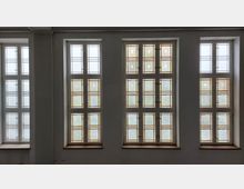 Drei große Fenster mit jeweils mehreren kleinen Sprossen und buntem Glas in geometrischen Mustern. Die Scheiben sind mit dezenten floralen Motiven verziert, und der Raum wirkt durch den Lichteinfall hell.