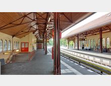 Bahnhof mit überdachten Bahnsteigen, roten Stahlträgern und bogenförmigen Fenstern. Auf beiden Seiten der Gleise befinden sich Wartebereiche mit Sitzbänken und Ticketautomaten; darüber digitale Anzeigetafeln.