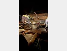 Große Werkstatt- oder Garagenhalle mit Holzdecke und Stahlträgern, in der mehrere Autos stehen und Werkbänke sowie Geräte im Hintergrund zu sehen sind. Im Vordergrund befindet sich ein Lounge-Bereich mit Sesseln, Couchtisch und Teppich auf Betonboden, seitlich hängen Poster und Lampen an den Wänden.