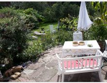 Ein Garten mit einer Terrasse aus Steinplatten, auf der eine weiße Sitzbank und ein mit einer Spitzen-Tischdecke bedeckter Tisch stehen. Im Hintergrund befinden sich Rasenflächen, Liegestühle, viele Pflanzen und Bäume sowie ein geschlossener Sonnenschirm.