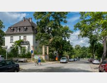 Straßenszene in einer grünen Wohngegend mit einem großen weißen Haus im historischen Stil, flankiert von Bäumen. Im Vordergrund verläuft eine gepflasterte Straße mit parkenden Autos und einer Person, die ein Fahrrad schiebt.