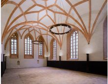Großer Raum mit gotischem Kreuzrippengewölbe und sichtbaren roten Backsteinrippen. Hohe, schmale Spitzbogenfenster mit dekorativem Maßwerk und modernen, ringförmigen Hängeleuchten dominieren die Gestaltung.