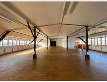 Großer, offener Raum mit Holzboden und weißer Decke, gestützt von Metallträgern. Entlang der Seiten befinden sich große Fenster mit Blick nach draußen, sowie Heizkörper unter den Fenstern.