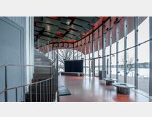 Moderner Innenraum mit großen bodentiefen Glasfenstern, die eine Aussicht auf eine winterliche Landschaft und ein Gewässer bieten. Der Raum hat eine gebogene, offene Treppe mit Metallgeländer, einen roten Boden und eine Holzdecke mit akustischen Elementen.