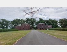 Zwei symmetrische Backsteingebäude mit mehreren Fenstern stehen links und rechts einer asphaltierten Straße, die zu einer großen Antennenanlage führt. Im Hintergrund sind hohe Bäume und ein bewölkter Himmel zu sehen, während der Bereich mit Rasen und Hecken eingefasst ist.