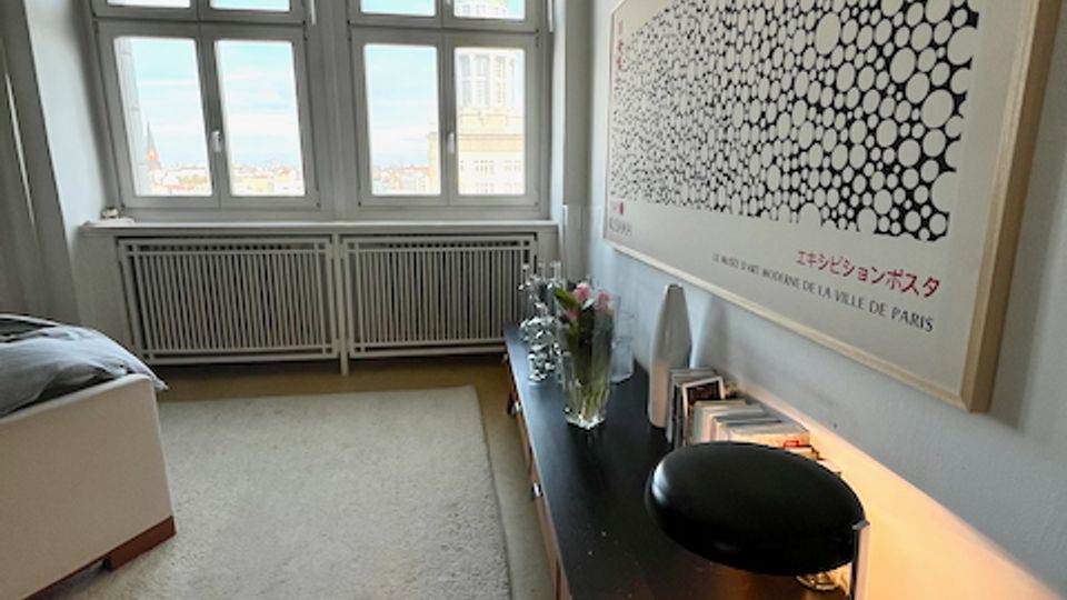 Schmales Schlafzimmer mit großen, mehrteiligen Fenstern und Heizkörper darunter, hellem Teppichboden und einem Bett am linken Rand. Entlang der rechten Wand steht eine lange Kommode mit Lampe und Dekoration, darüber hängt ein großes gerahmtes Poster.