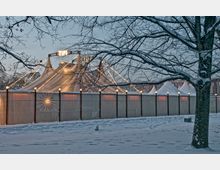 Ein großes weißes Zelt mit Lichterketten auf den Zeltspitzen, umgeben von einem hölzernen Lattenzaun. Im Vordergrund liegt eine schneebedeckte Landschaft mit einem kahlen Baum.