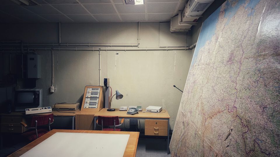 Kleiner, fensterloser Arbeitsraum mit niedriger Decke, Neonleuchte, sichtbaren Leitungen und einem langen Schreibtisch mit alten Kommunikationsgeräten und Telefon. Rechts lehnt eine große, detaillierte Landkarte an der Wand; vorne steht ein großer Arbeitstisch.