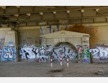 Unterseite einer Brücke aus Metallträgern mit einer Wand aus Stein, auf der zahlreiche bunte Graffiti gesprüht wurden. Im Vordergrund sind sandiger Boden und drei rot-weiß gestreifte Absperrpfosten zu sehen.