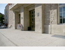 Detailansicht eines massiven sandsteinfarbenen Gebäudeeingangs mit breiter Holztür im historischen Stil. Im Vordergrund steht auf dem gepflasterten Vorplatz ein Informationsaufsteller; im Hintergrund sind weitere Gebäude und ein Straßenschild sichtbar.