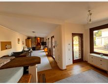 Langer Wohnraum mit Holzboden, großen Fenstern und einer Glastür, die nach außen führt. Der Raum ist möbliert mit einer Sofaecke, einem Holzschrank und einer Kommode; die Wände sind hell gestrichen, mit einigen Pinnwänden dekoriert.