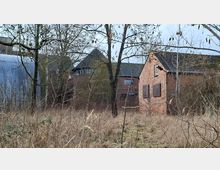 Verwildertes Gelände mit hohem Gras und kahlen Bäumen vor mehreren Backsteingebäuden mit dunklen Ziegeldächern, darunter ein runder Turmanbau mit Fensterband. Links steht eine große Halle mit Wellblechverkleidung, rechts ein kleines Backsteinhaus mit teilweise vernagelten Fenstern.