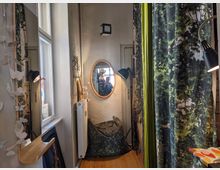 Ein kleiner Raum mit hellem Holzboden und weißen Wänden, dekoriert mit einem ovalen Spiegel an der Wand, flankiert von schwarzen Stehlampen. Rechts hängen grüne Vorhänge mit Blättermuster, links ein Fenster und eine schmale Holzkonsole mit Papierdekorationen.