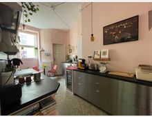 Kleine Küche mit rosa Wänden und einem Mix aus modernen und Vintage-Elementen. Links befindet sich eine schwarze Arbeitsplatte mit Spüle, rechts eine Edelstahlkommode mit Küchenutensilien, und im Hintergrund steht ein runder Esstisch mit roten Stühlen vor einem großen Fenster.