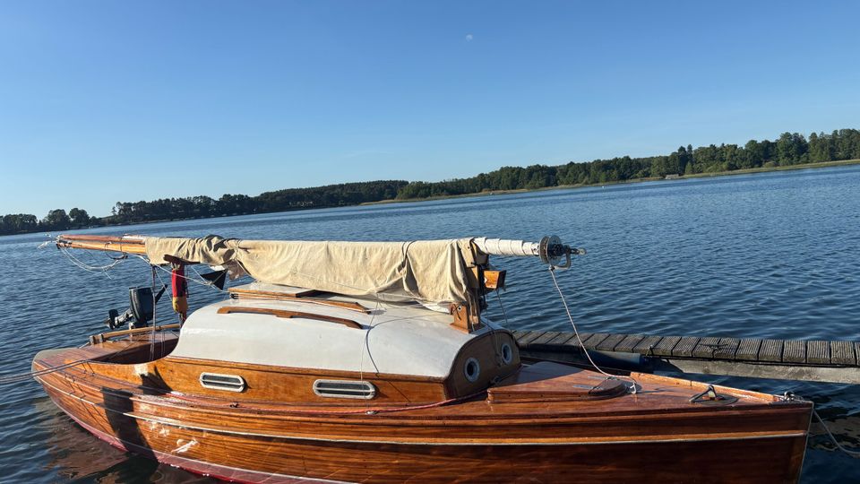 Kleines hölzernes Segelboot mit aufgerolltem, beigefarbenem Segel liegt an einem Steg auf einem ruhigen See. Im Hintergrund sind eine flache, bewaldete Uferlinie und ein klarer blauer Himmel zu sehen.