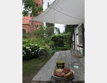 Ein Gartentisch mit einem Teller Essen, einer Tasse und einem Krug Wasser steht unter einem weißen Sonnensegel. Im Hintergrund sind ein gepflegter Garten mit Blumen, ein Fahrrad und eine Kirche mit rotem Ziegelmauerwerk zu sehen.