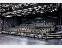 Innenraum eines kleinen schwarzen Theater- oder Veranstaltungssaals mit ansteigenden Reihen grauer Stühle auf Tribünen. Über der Sitzfläche hängt ein Metallträger-Rigg mit Bühnenbeleuchtung und Technik, daneben seitliche Treppenaufgänge.