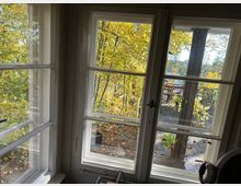 Blick durch große weiße Sprossenfenster auf eine herbstliche Terrasse mit Holzfußboden, buntem Laub an den Bäumen und einer Sitzgruppe im Hintergrund. Im Vordergrund sind Fensterrahmen und ein Teil des Innenraums zu sehen.