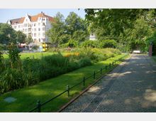 Ein kleiner Stadtpark mit gepflastertem Weg, umgeben von grünen Bäumen, Blumenbeeten und Rasenflächen. Im Hintergrund sind ein historisches, weißes Gebäude mit roten Ziegeldächern und eine Bushaltestelle zu sehen.