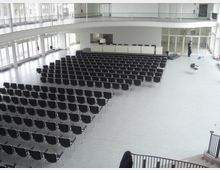 Ein heller Veranstaltungsraum mit Reihen schwarzer Stühle, einer erhöhten Bühne mit einem Rednerpult im Hintergrund und einer umlaufenden Galerie. Große Fensterfronten sorgen für viel Tageslicht.