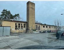 Ehemaliges Industriegebäude aus gelben Backsteinen mit einem hohen Schornstein in der Mitte. Die Fenster sind teilweise beschädigt, und ein Bauzaun umgibt das Gelände, das Graffiti und überwuchertes Gelände zeigt.