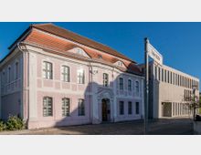 Rosa-weißes historisches Gebäude mit Sprossenfenstern und verziertem Eingang zur rechten Straßenseite, Beschriftung „Kleist-Museum“ über dem Tor; daneben ein moderner Betonbau mit schmalen vertikalen Fenstern entlang der Fassade. Im Vordergrund ein Straßenschild mit der Aufschrift „Faberstraße“.