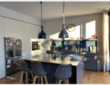 Moderne Küche mit Kochinsel, Hockern und großer Arbeitsplatte. Im Hintergrund befinden sich Einbauschränke, ein Einbauofen, ein Kühlschrank mit Magneten sowie Hängelampen und ein Fenster, das natürliches Licht hereinlässt.